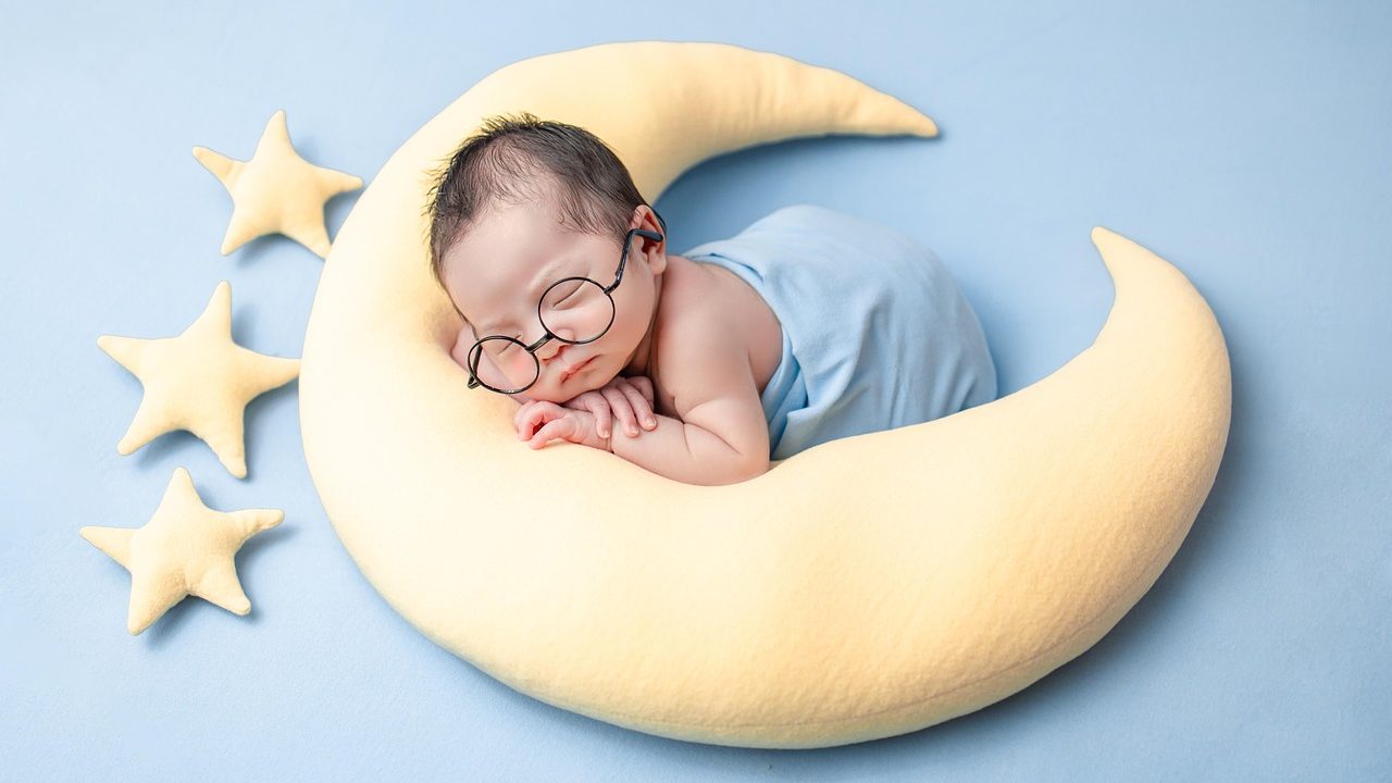 Parent sitting at a desk with paperwork and laptop next to a sleeping newborn in a bassinet.