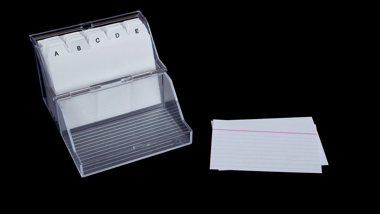 A clean wooden desk featuring a stack of white index cards, a black pen, and a small card box.