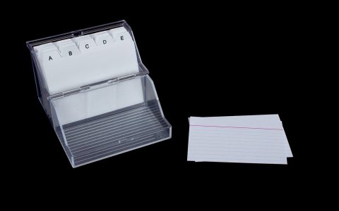 A clean wooden desk featuring a stack of white index cards, a black pen, and a small card box.