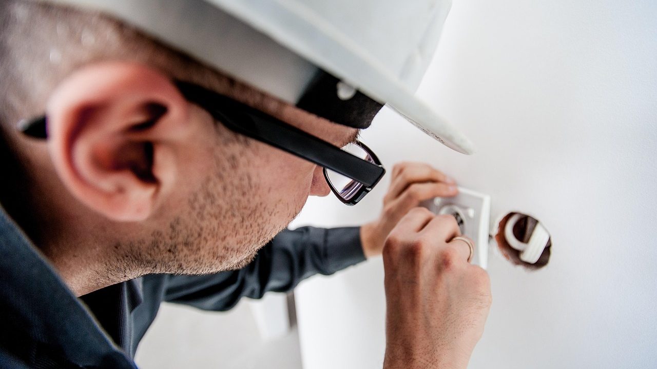 An electrician examining a storm-damaged overhead electric meter socket and service mast on a house exterior.