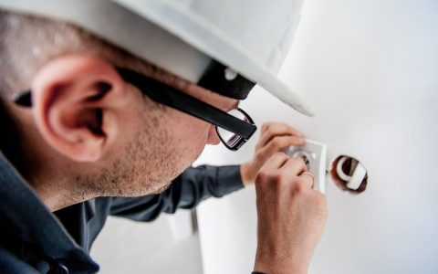 An electrician examining a storm-damaged overhead electric meter socket and service mast on a house exterior.