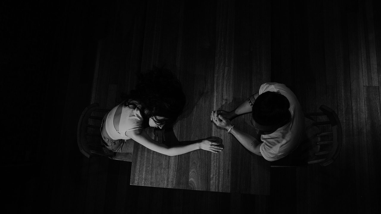 An engaged couple sitting at a wooden table reviewing financial documents together on a laptop in a brightly lit room.