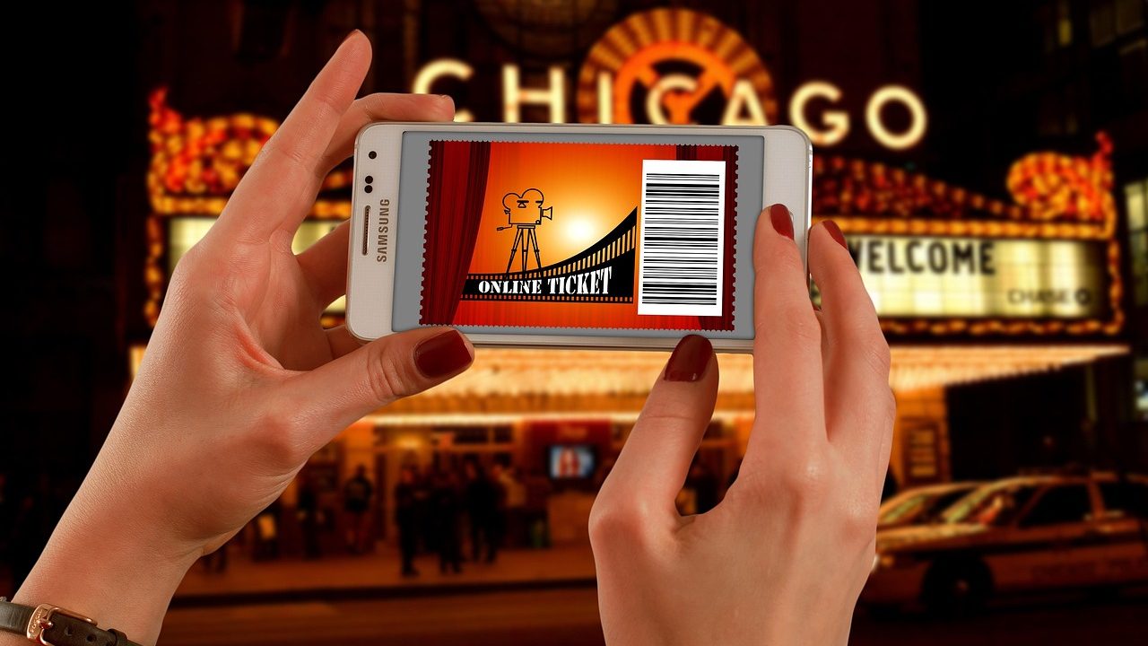 A person holding a smartphone with a digital ticket barcode outside a concert arena entrance at dusk.