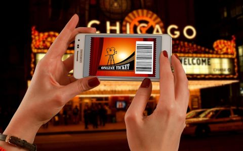 A person holding a smartphone with a digital ticket barcode outside a concert arena entrance at dusk.