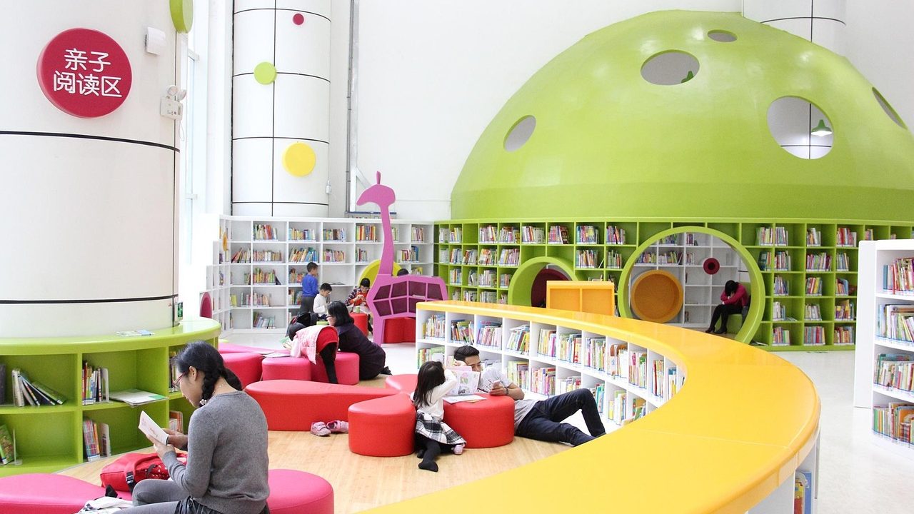Young child sitting on a library floor reading a large picture book.