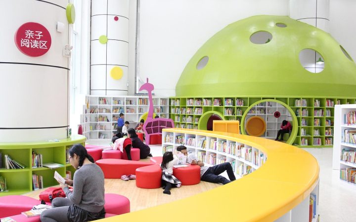 Young child sitting on a library floor reading a large picture book.