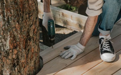 Close-up of a person applying protective stain to a weathered wooden deck board with a brush.