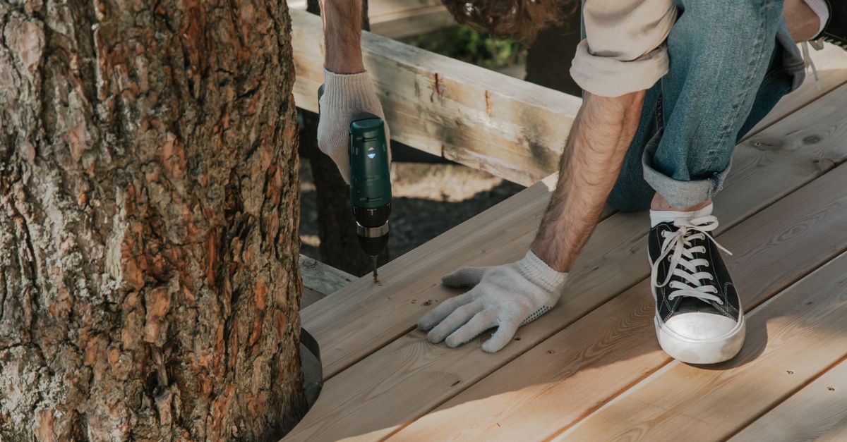 Close-up of a person applying protective stain to a weathered wooden deck board with a brush.