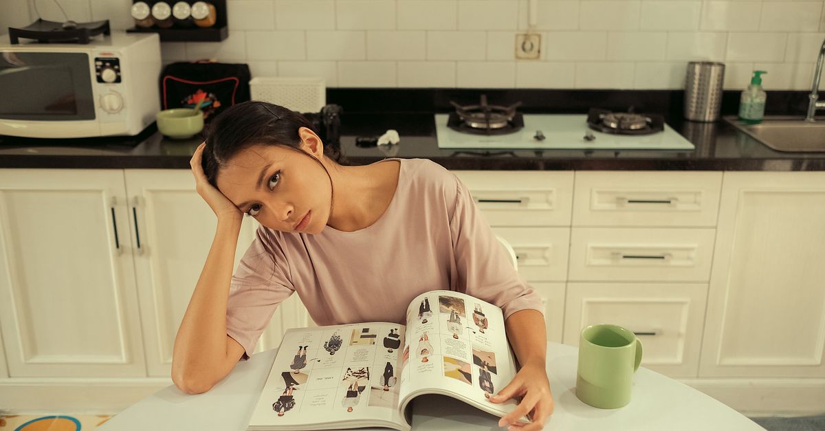 A woman looking tired and stressed while sitting at a kitchen table with papers.