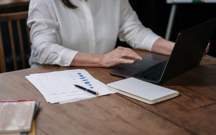A laptop open to a spreadsheet on a wooden desk with a calculator and notepad.