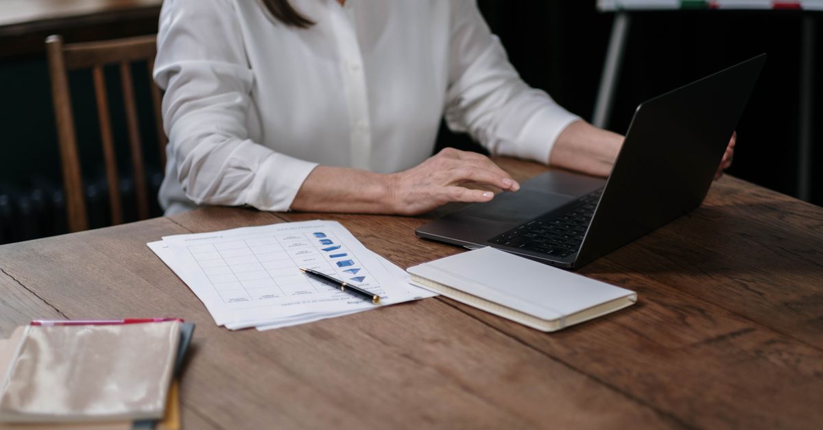 A laptop open to a spreadsheet on a wooden desk with a calculator and notepad.