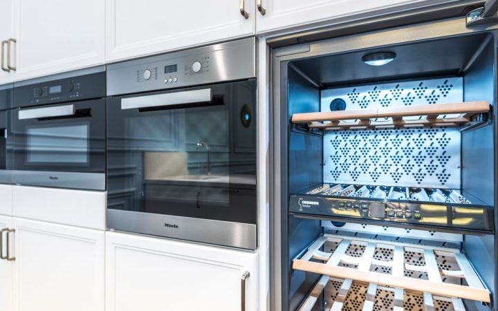 A stainless steel built-in wine refrigerator installed in a modern kitchen with wooden shelves holding wine bottles.