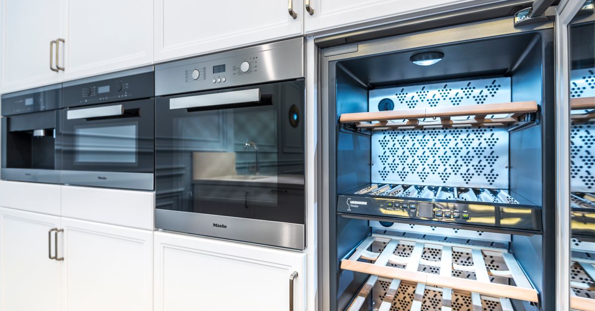 A stainless steel built-in wine refrigerator installed in a modern kitchen with wooden shelves holding wine bottles.